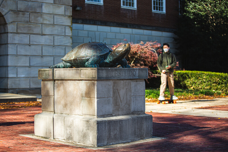 View of Testudo statue in front of McKeldin Library with fall foliage and a student walking by wearing a mask.